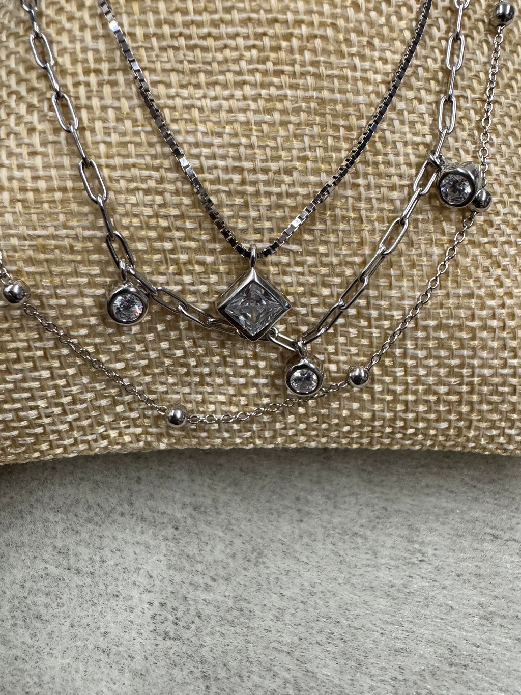 The sterling silver layered necklace arranged across woven cream fabric, the three strands sweeping past the square cubic zirconia and two round bezel-set stones.