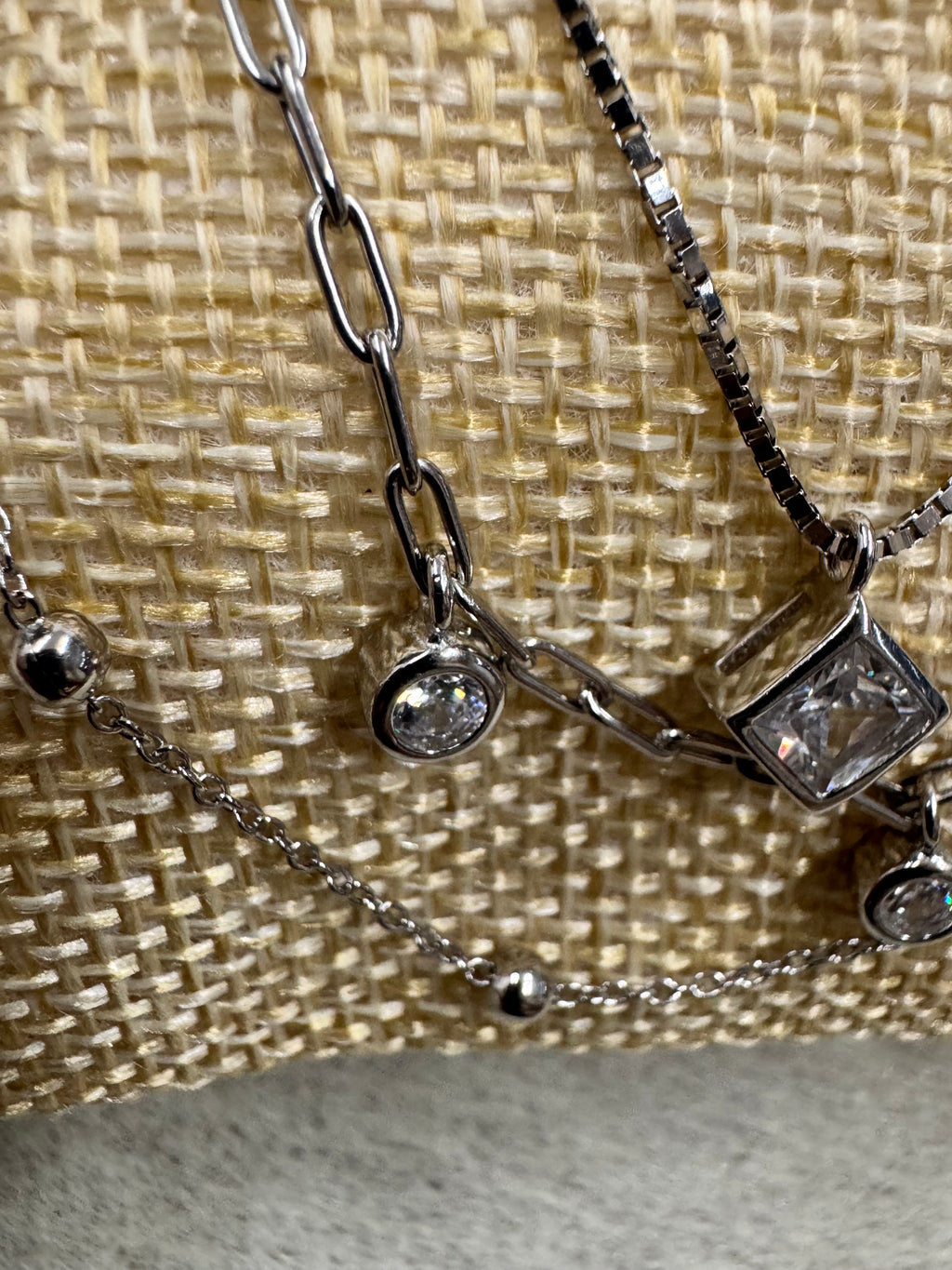 Close view on cream woven fabric of the sterling silver necklace, with the square cubic zirconia above a round bezel-set stone on the paperclip chain and the satellite bead chain to the right.
