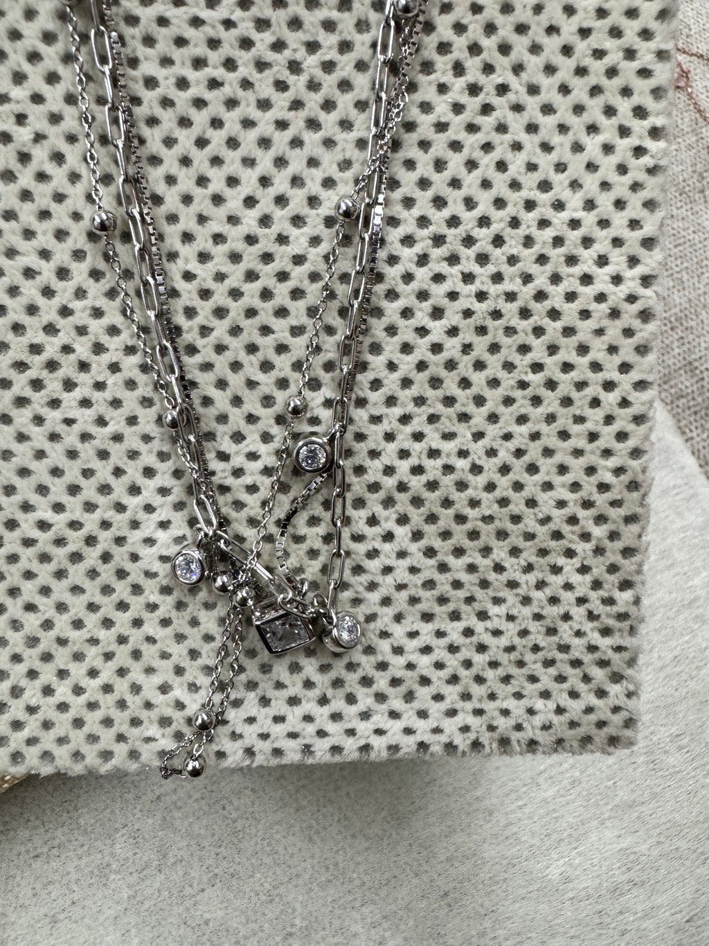 The sterling silver layered necklace lying diagonally across cream fabric with grey dots, the square cubic zirconia and two round drops gathered near the centre.
