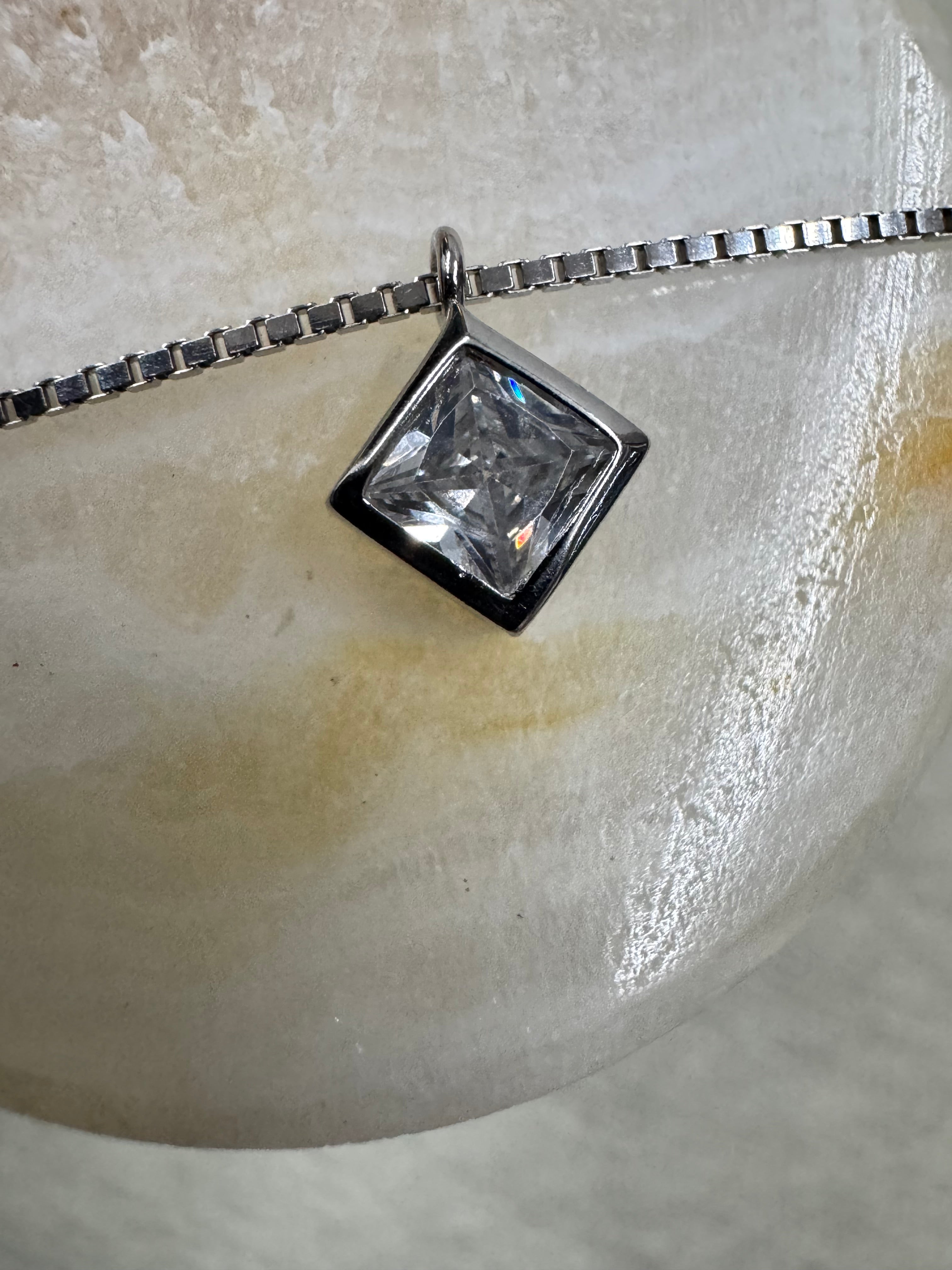 The square bezel-set cubic zirconia and box chain of the sterling silver necklace resting on a pale polished stone prop.