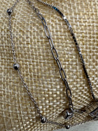 Detail of the box, paperclip and satellite chains of the sterling silver necklace on cream woven fabric, with the polished silver balls and one bezel-set cubic zirconia visible.