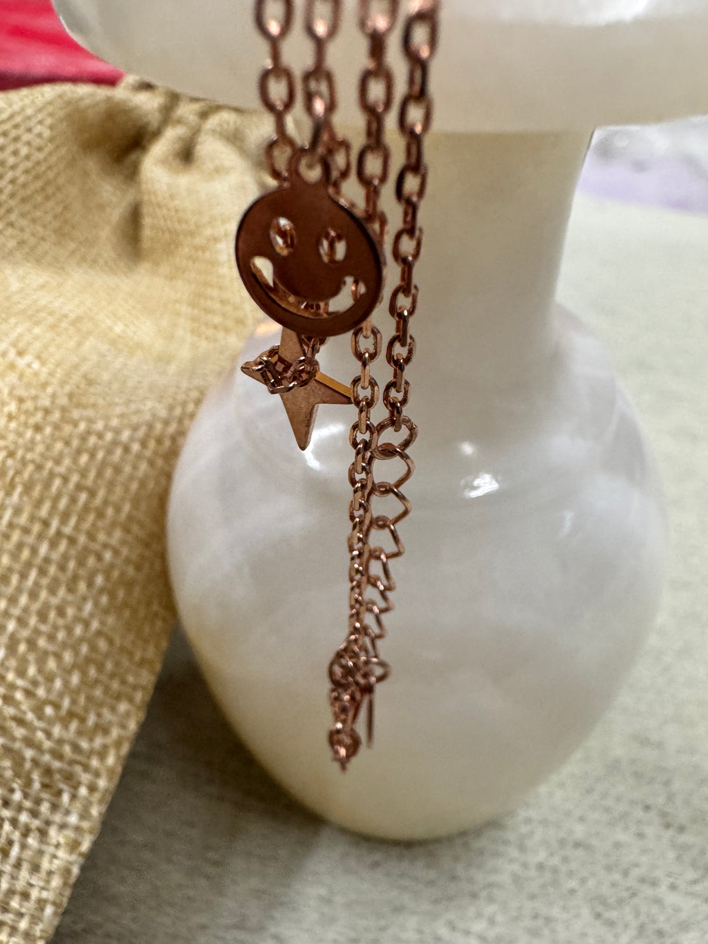 The rose-tone sterling silver necklace draped over a white stone ornament beside a jute pouch, a smiley disc and star in view.