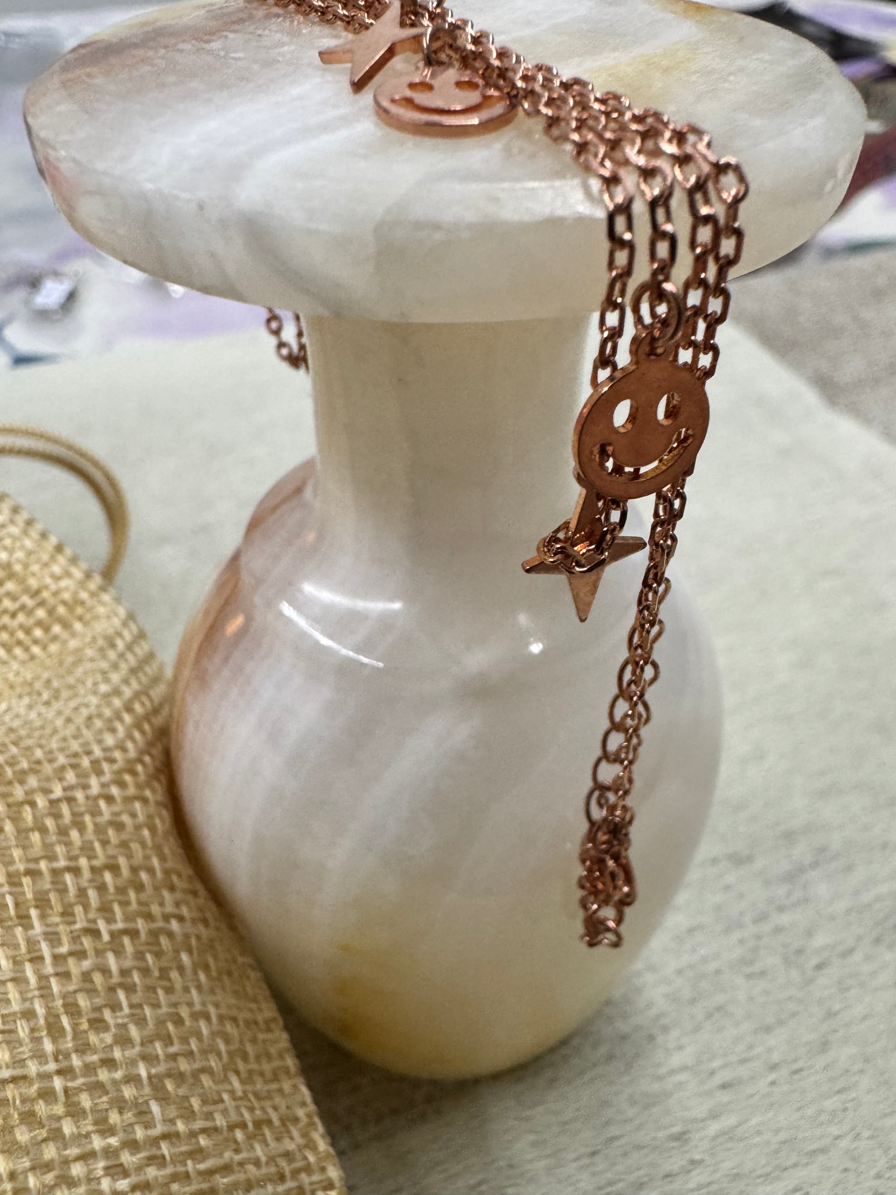 Wider view of the rose-tone sterling silver station necklace over a white stone ornament, several smiley discs and stars along the chain.