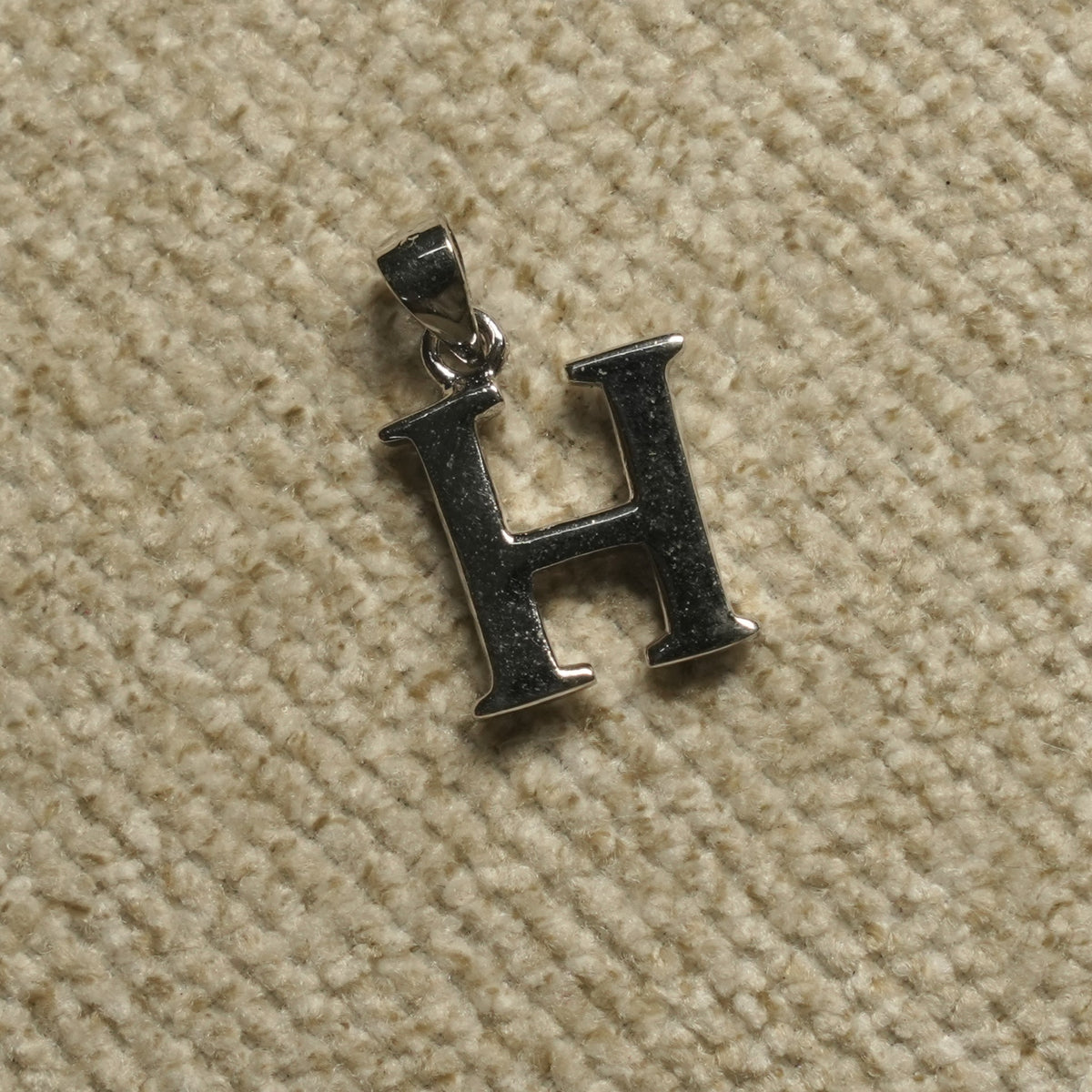 Polished sterling silver initial H pendant with hooked terminals, resting on pale grey felt.