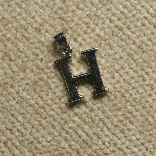 Polished sterling silver initial H pendant with hooked terminals, resting on pale grey felt.
