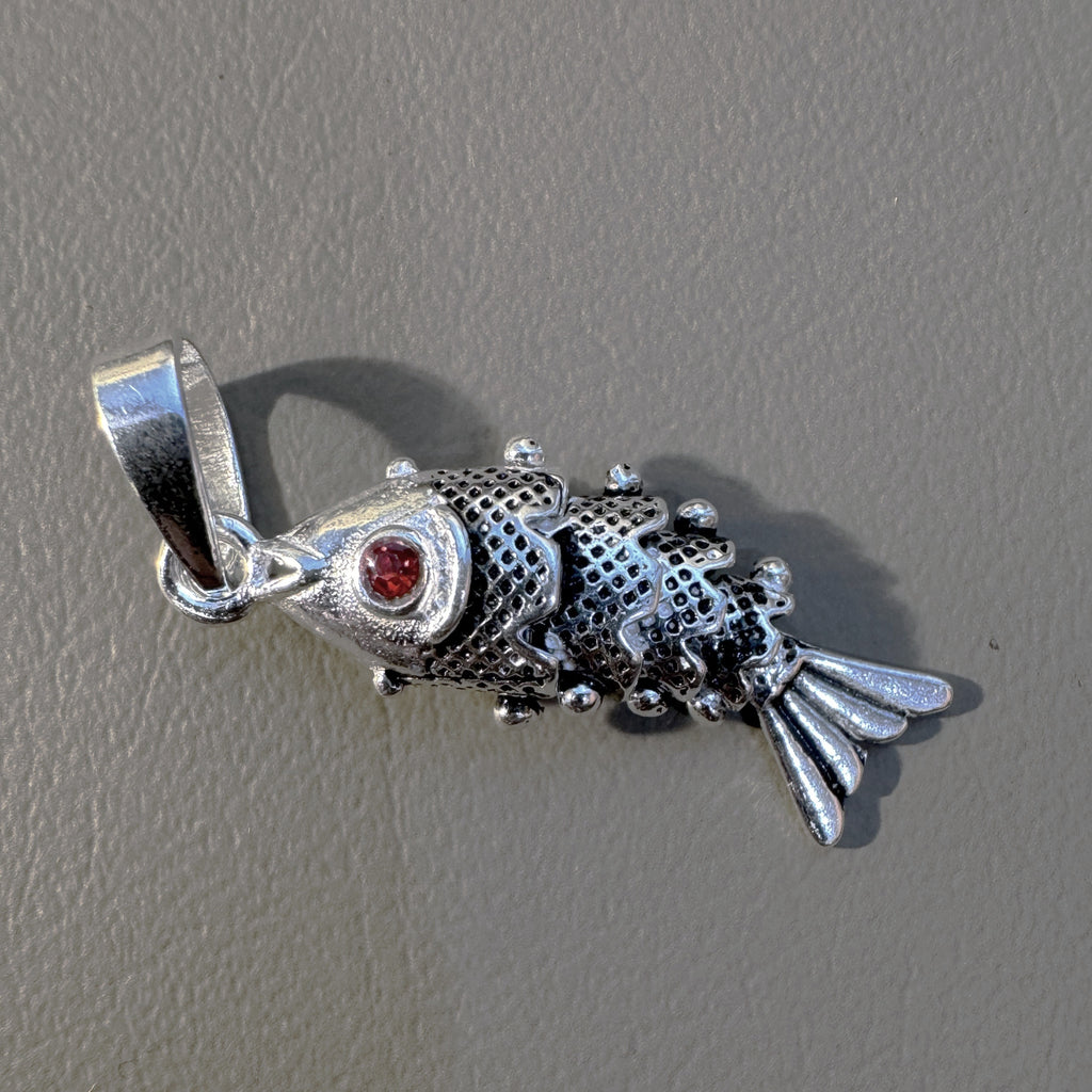 The jointed silver fish pendant lying on a grey surface with its bail to the left.
