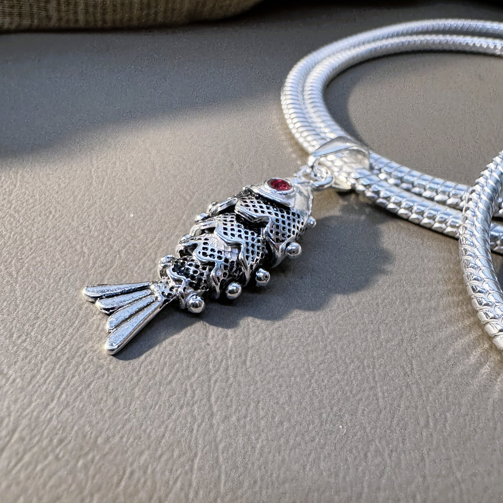 The oxidised fish pendant threaded on a snake chain, seen at an angle.