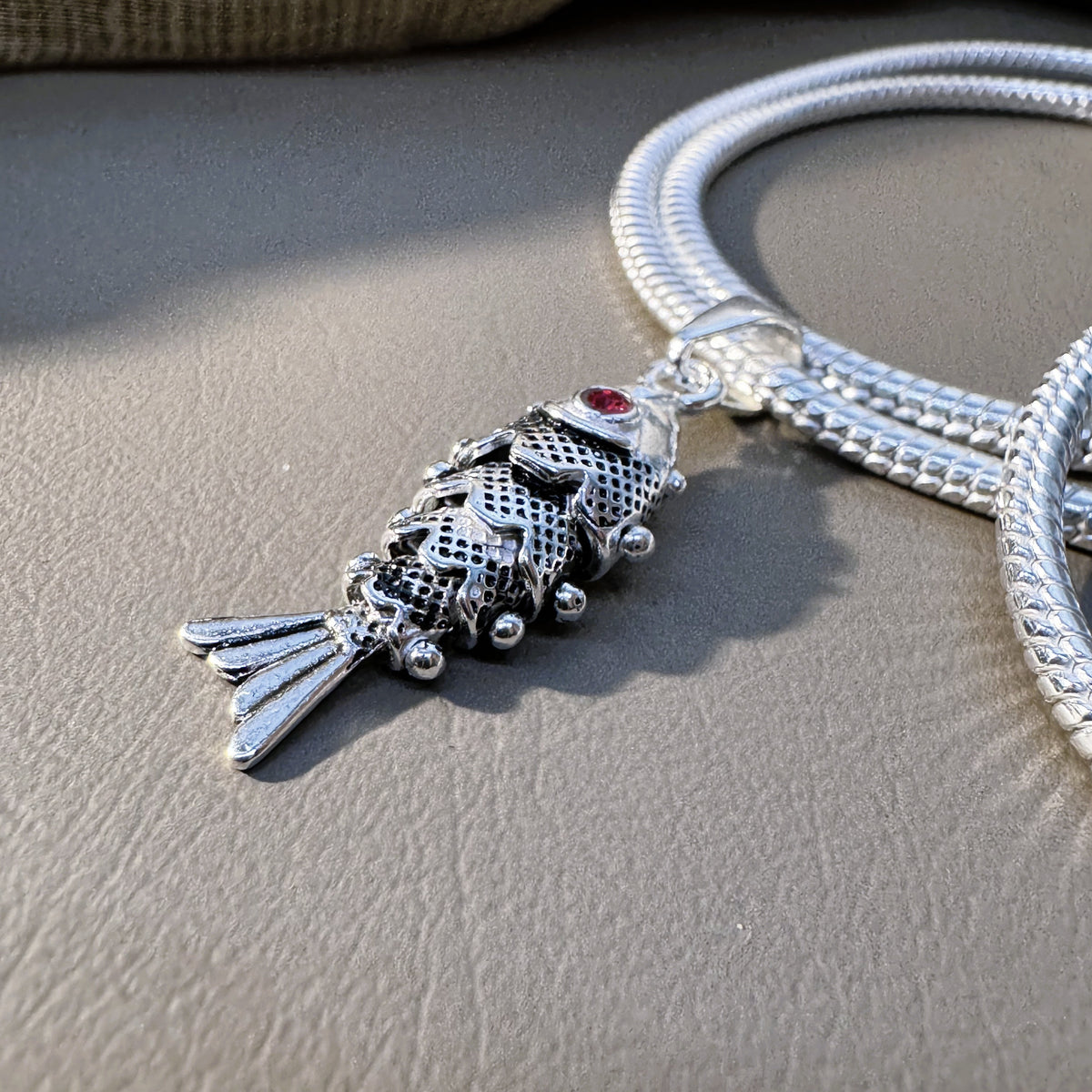 The oxidised fish pendant threaded on a snake chain, seen at an angle.