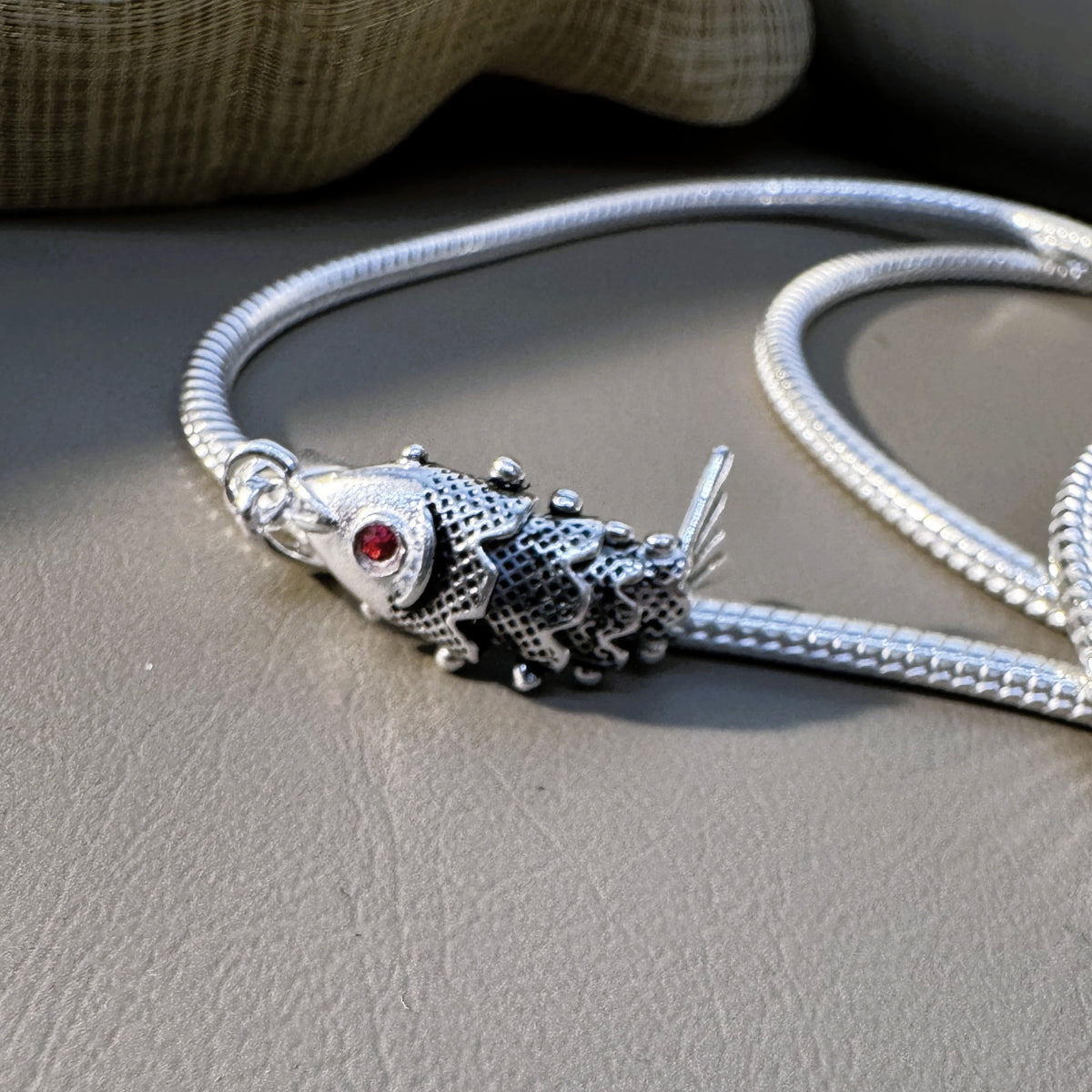 The silver fish pendant and chain lying together on a grey surface.