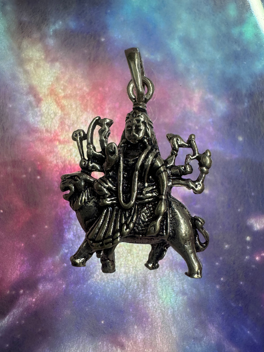 Oxidised sterling silver Goddess Durga pendant seated on her lion, against an iridescent nebula-print backdrop.