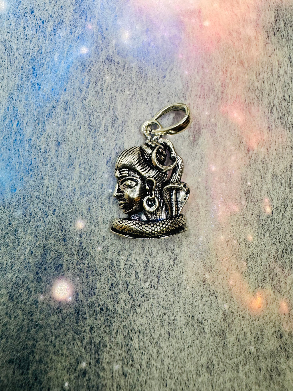 The sterling silver Shiva profile pendant on a grey and pink iridescent backdrop, bail at the top.