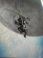 Oxidised sterling silver Ganesha pendant cast in a dancing pose, the trunk curled over a modak, on pale textured fabric in raking sunlight.