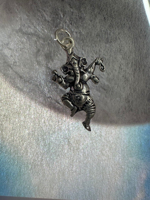 Oxidised sterling silver Ganesha pendant cast in a dancing pose, the trunk curled over a modak, on pale textured fabric in raking sunlight.