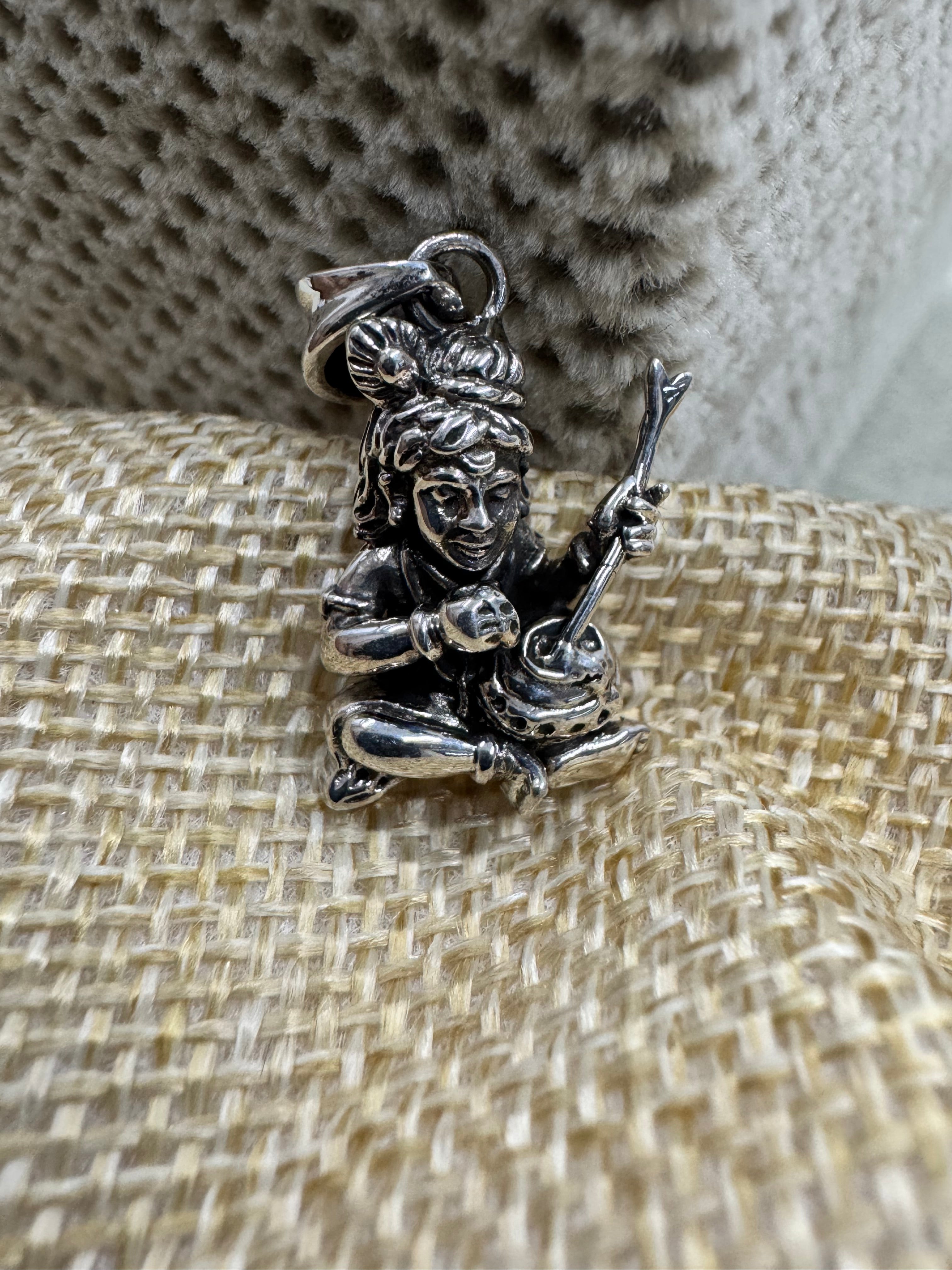 Sterling silver Bal Gopal pendant with churning stick, resting on woven beige jute beside grey velvet.