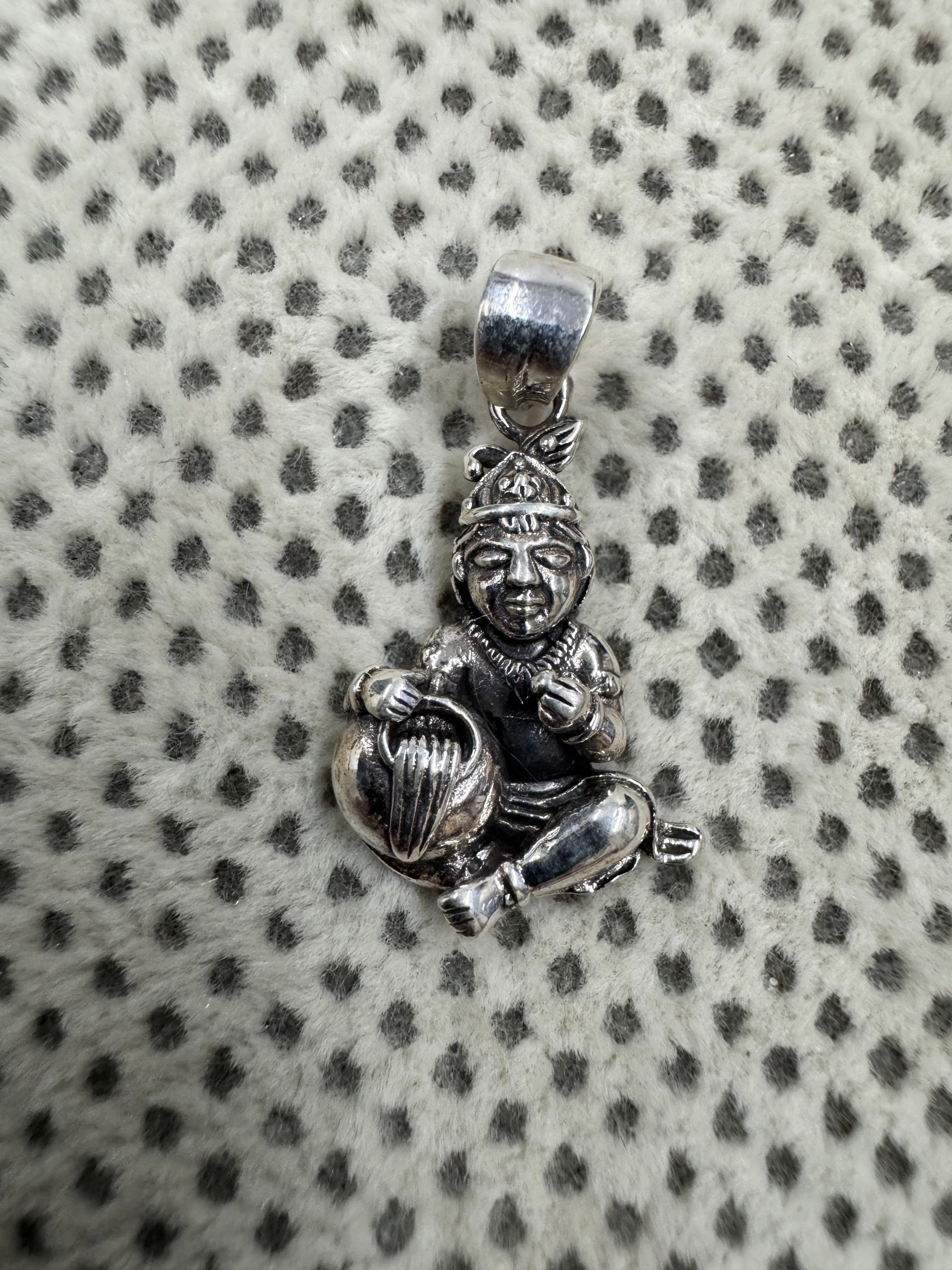 Cast sterling silver Bal Gopal pendant lying on grey dotted fabric, the round butter pot held under one arm.