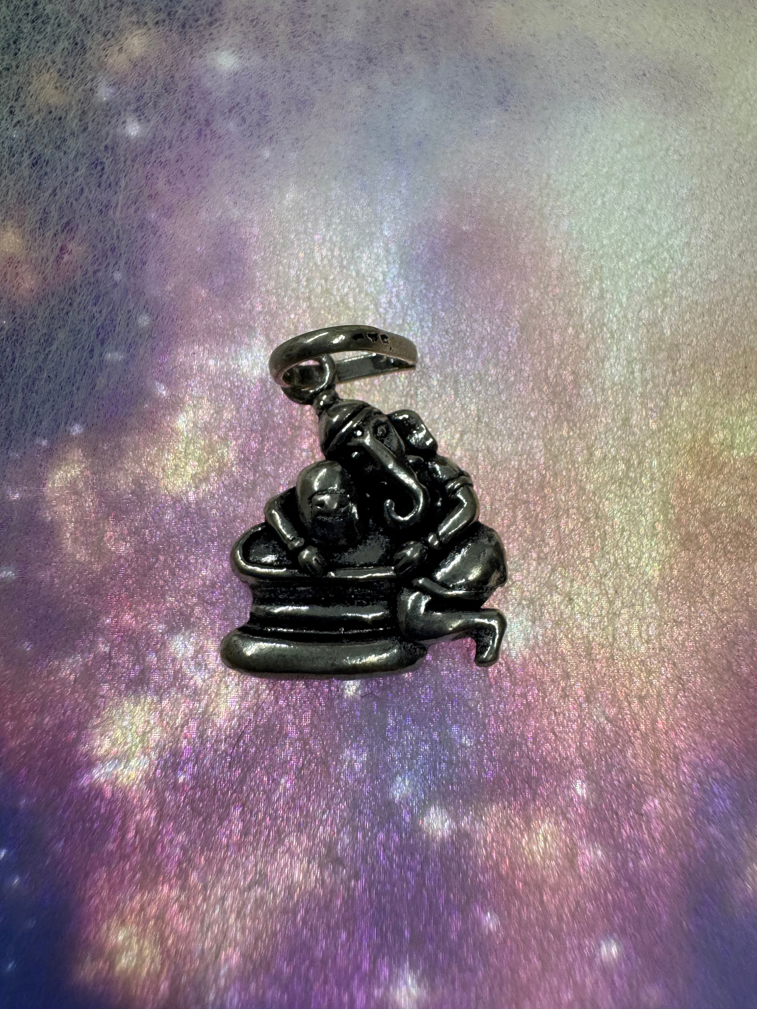 Sterling silver Ganesha pendant in a dark antique finish, seated on its stepped plinth against an iridescent pastel backdrop.