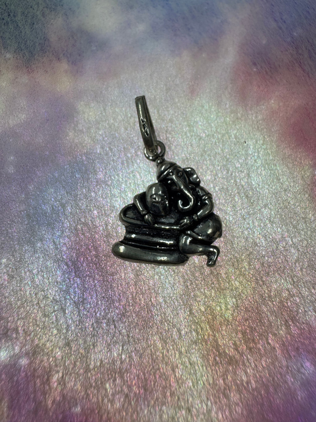 Antique-finish sterling silver Ganesha pendant seen from above on an iridescent surface, the trunk and ears in relief.