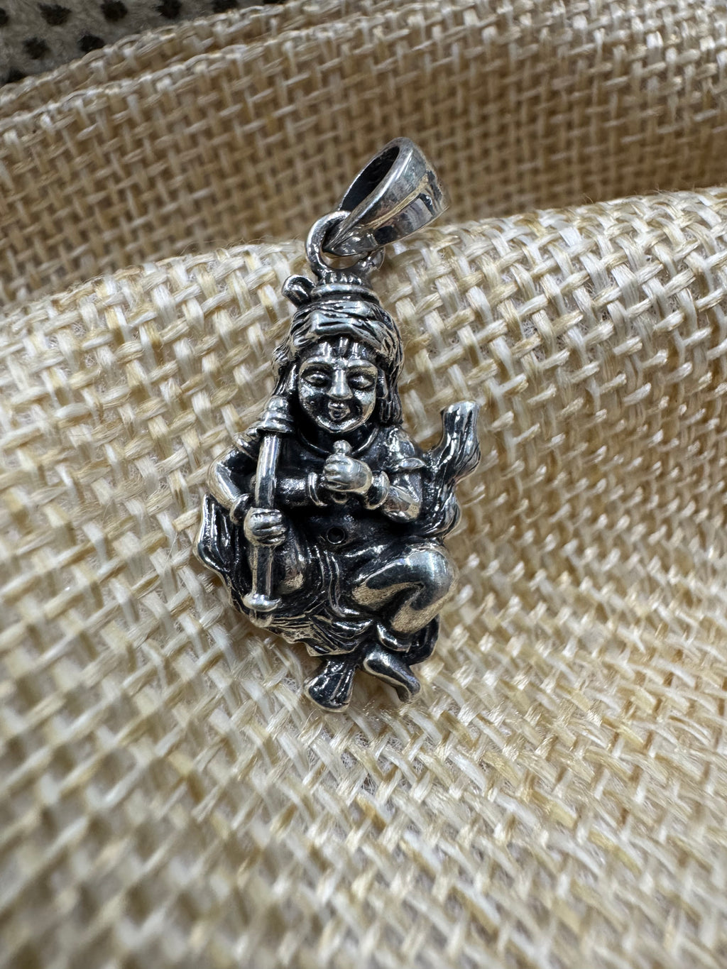 Sterling silver krishan pendant seated with a hearding stick, resting on a woven jute backdrop.