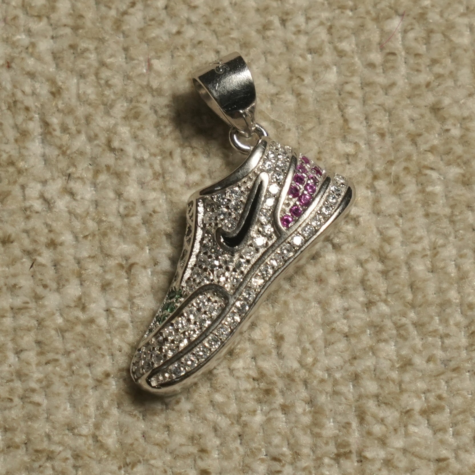 Sterling silver high-top trainer Pendant Bejewelled-set with cubic zirconia, with blush-pink and green cubic zirconia accents and a contrast black motif, on a beige woven backdrop.