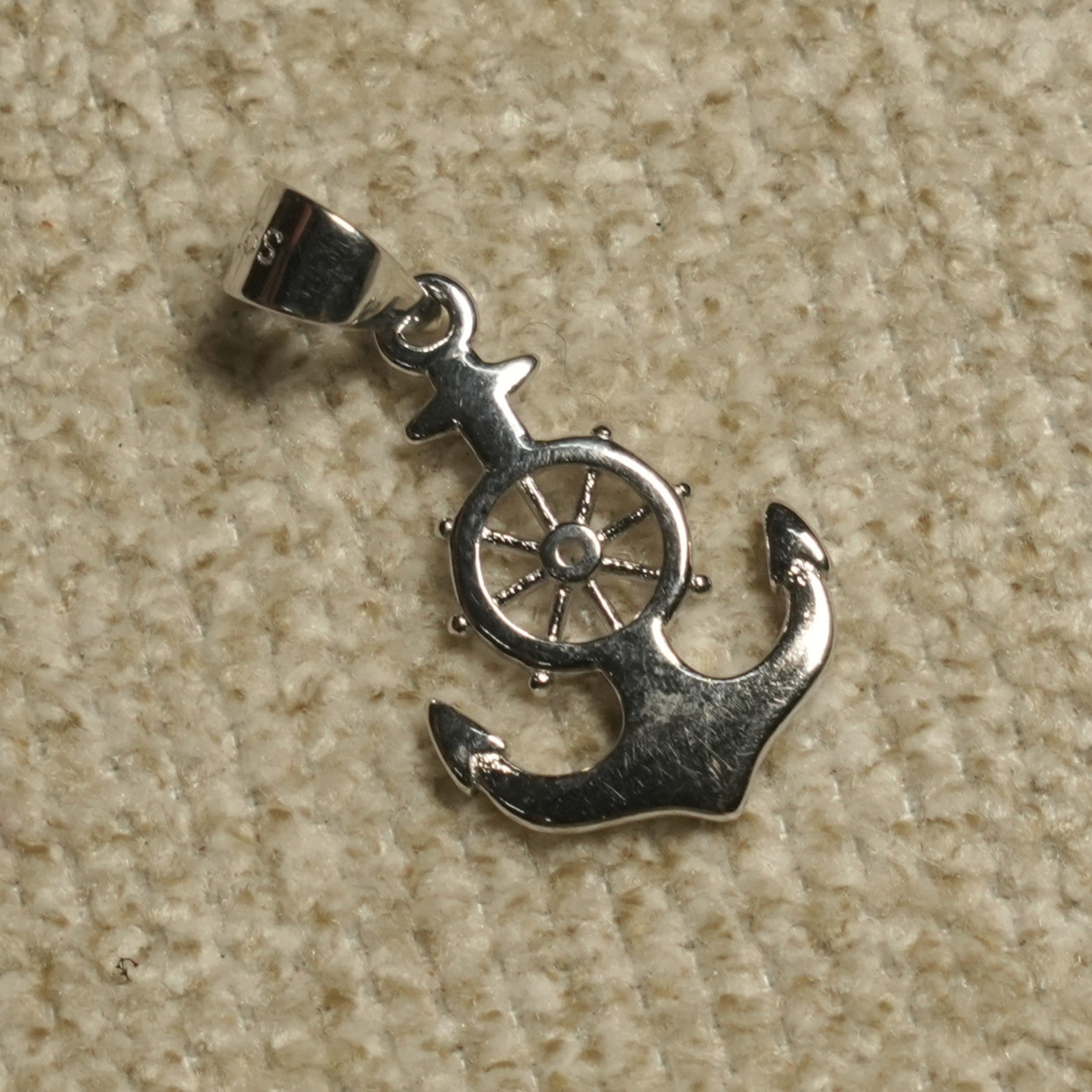 Polished sterling silver Pendant of an anchor crossed with a ship's wheel, resting on a beige woven backdrop.