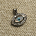 Sterling silver evil eye Pendant with a pale iridescent eye centre and a border of cubic zirconia, photographed on beige woven fabric.