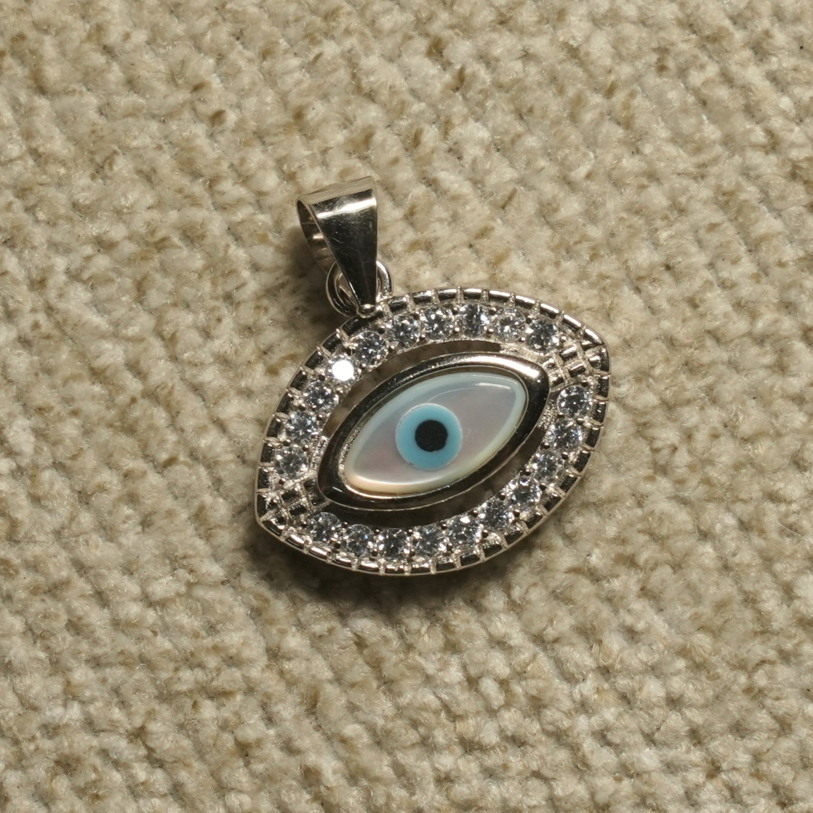 Sterling silver evil eye Pendant with a pale iridescent eye centre and a border of cubic zirconia, photographed on beige woven fabric.