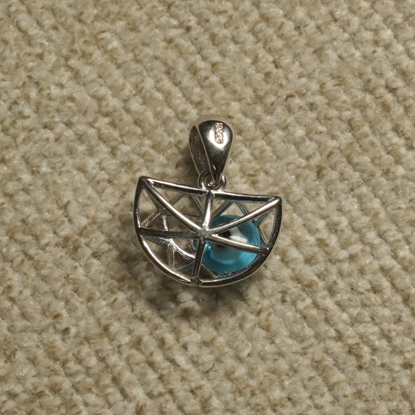 Sterling silver half-moon openwork cage Pendant holding a turquoise-blue eye bead, photographed on beige woven fabric.