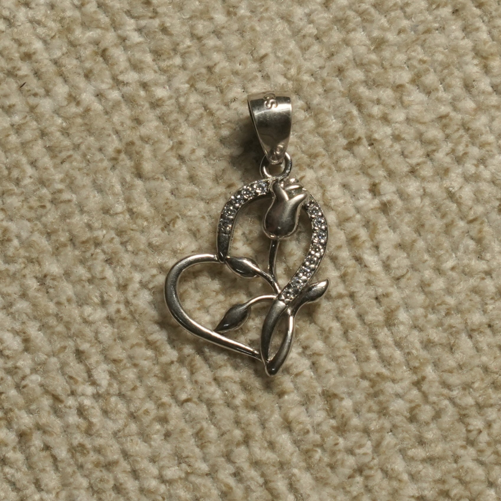 Sterling silver open heart Pendant with a sculpted rose motif and a line of cubic zirconia along one edge, photographed on beige woven fabric.