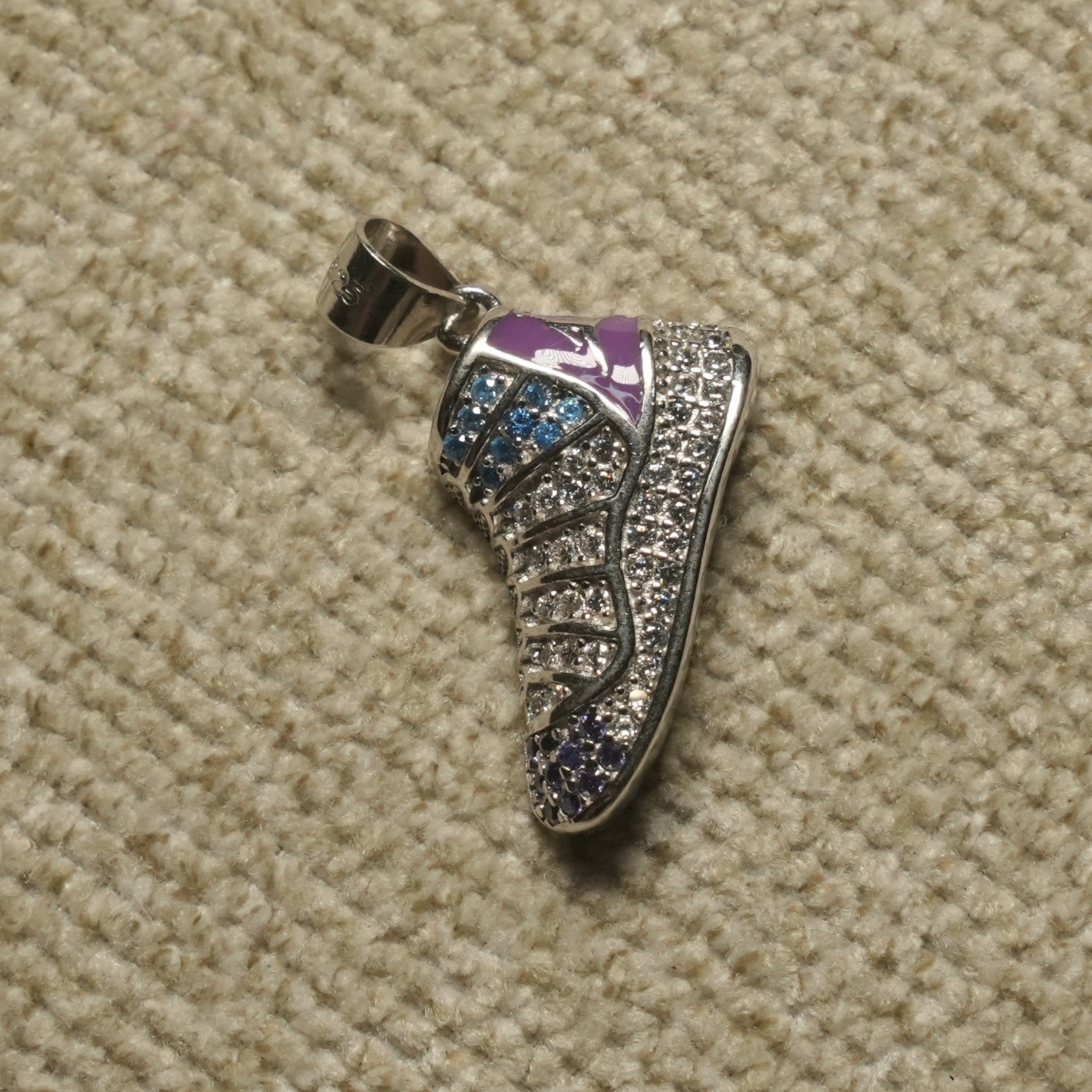 Sterling silver sneaker Pendant covered in clear, blue and violet cubic zirconia with a lilac enamel collar, photographed on beige woven fabric.