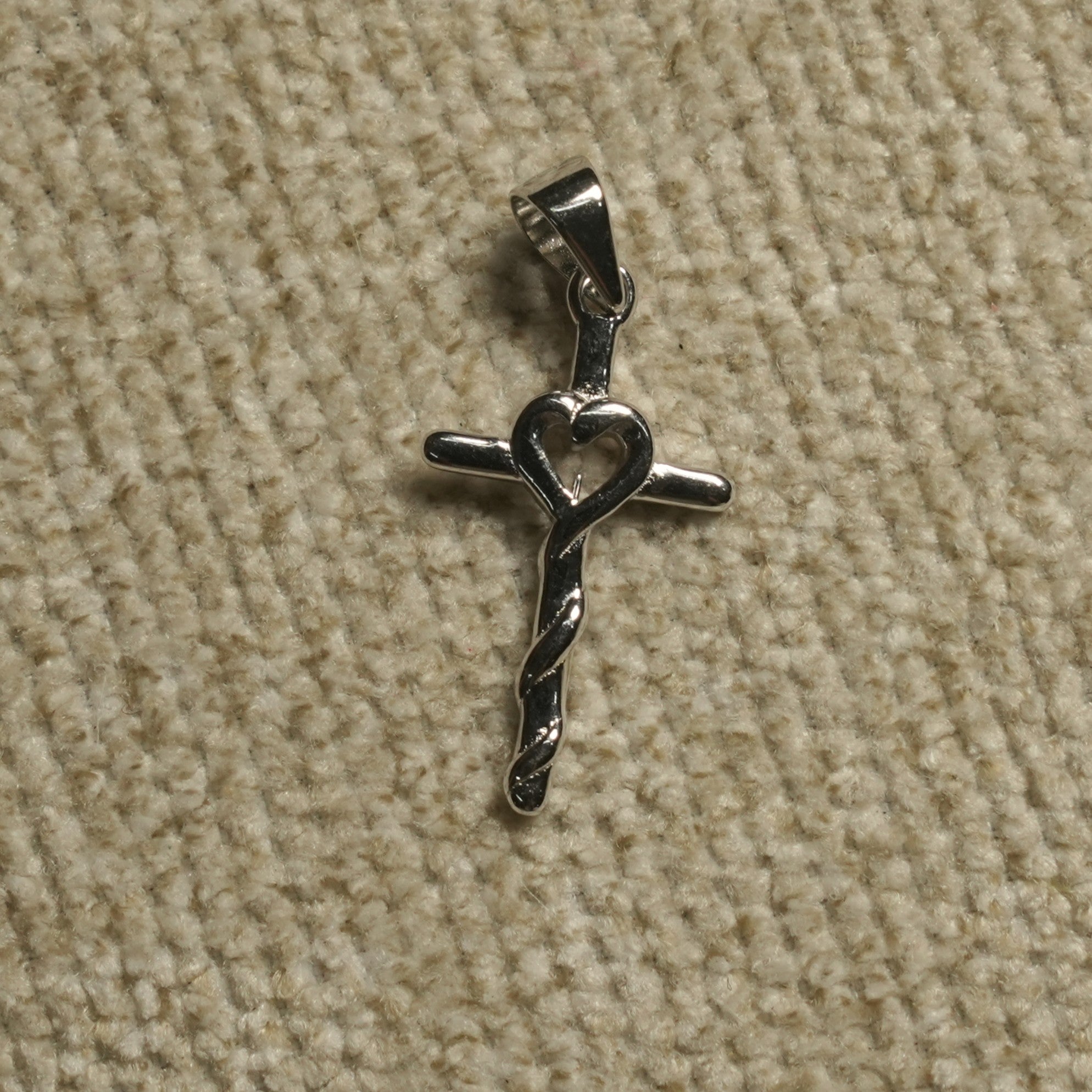 Polished sterling silver cross Pendant with an open heart at the centre and a twisted lower stem, photographed on beige woven fabric.