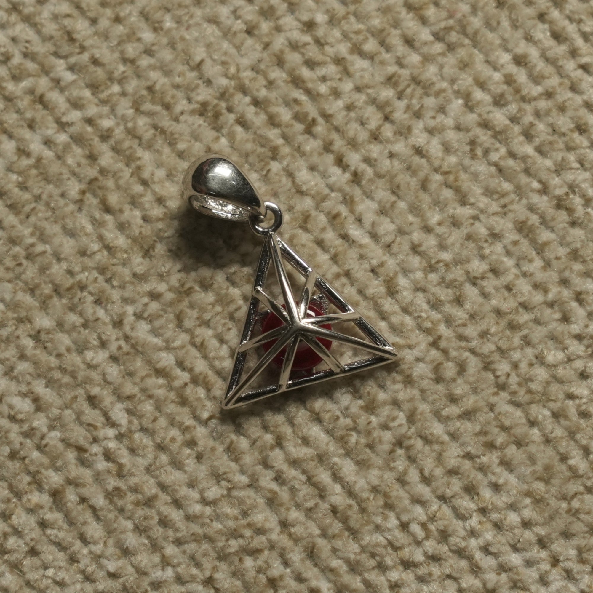 Sterling silver openwork triangle Pendant with a red bead at its centre, photographed flat on beige woven fabric.
