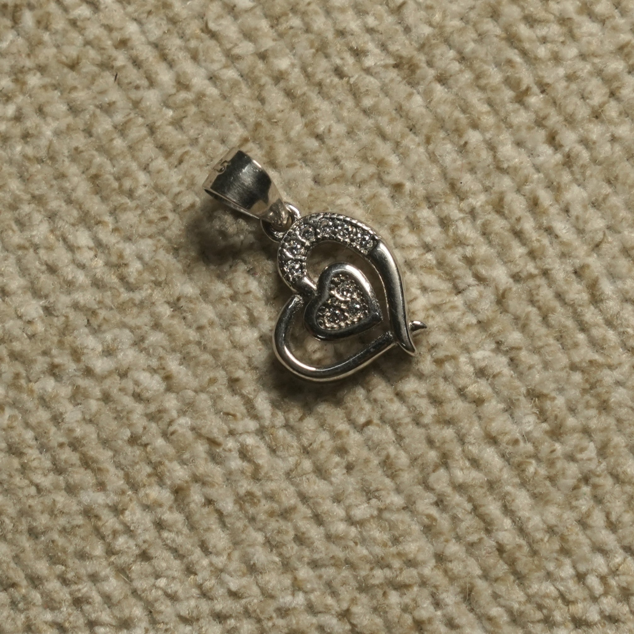 Sterling silver Pendant of an open heart holding a smaller Bejewelled cubic zirconia heart, photographed on beige woven fabric.