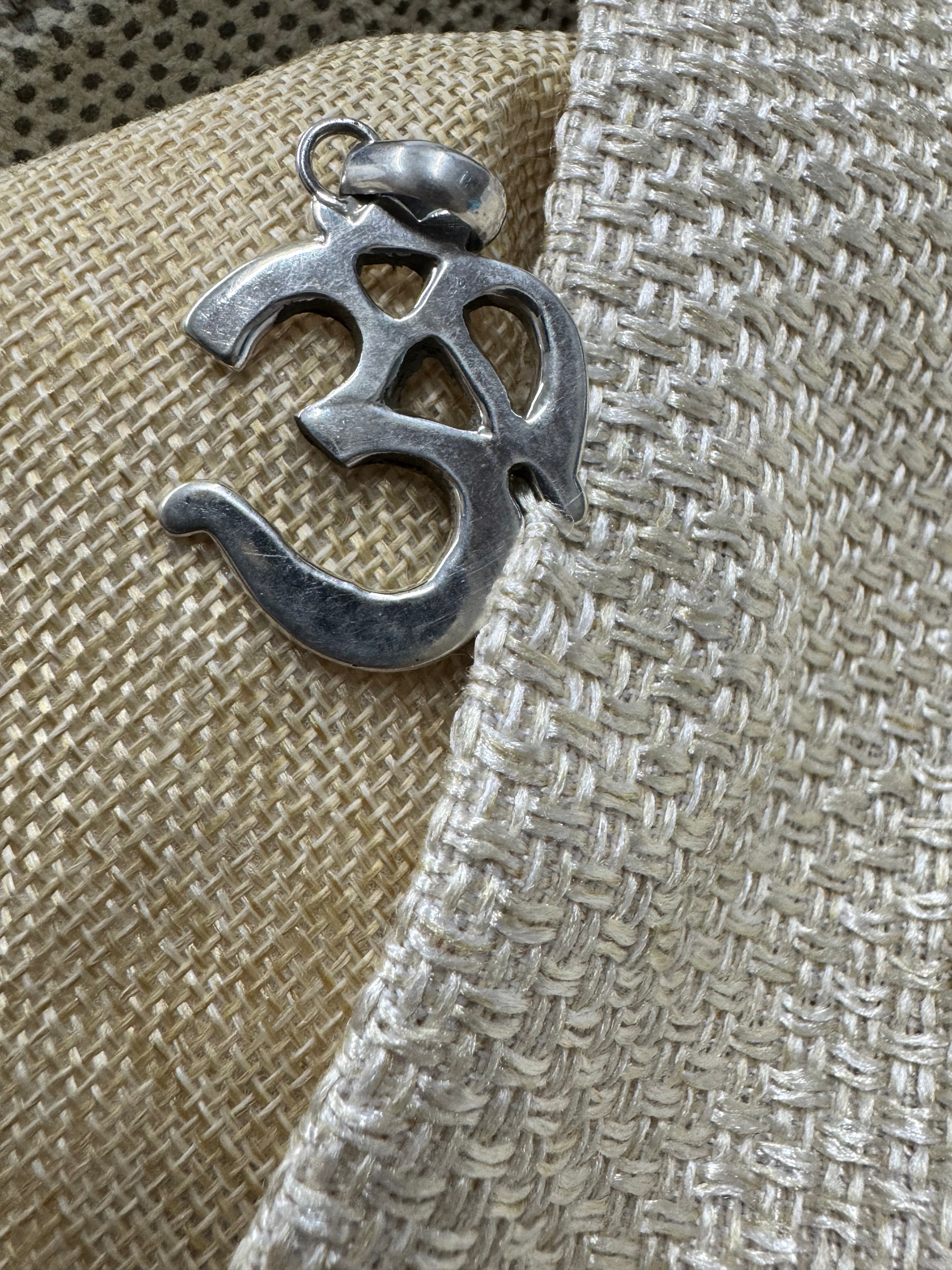 Polished sterling silver Om pendant with openwork curves, resting against woven beige fabric.