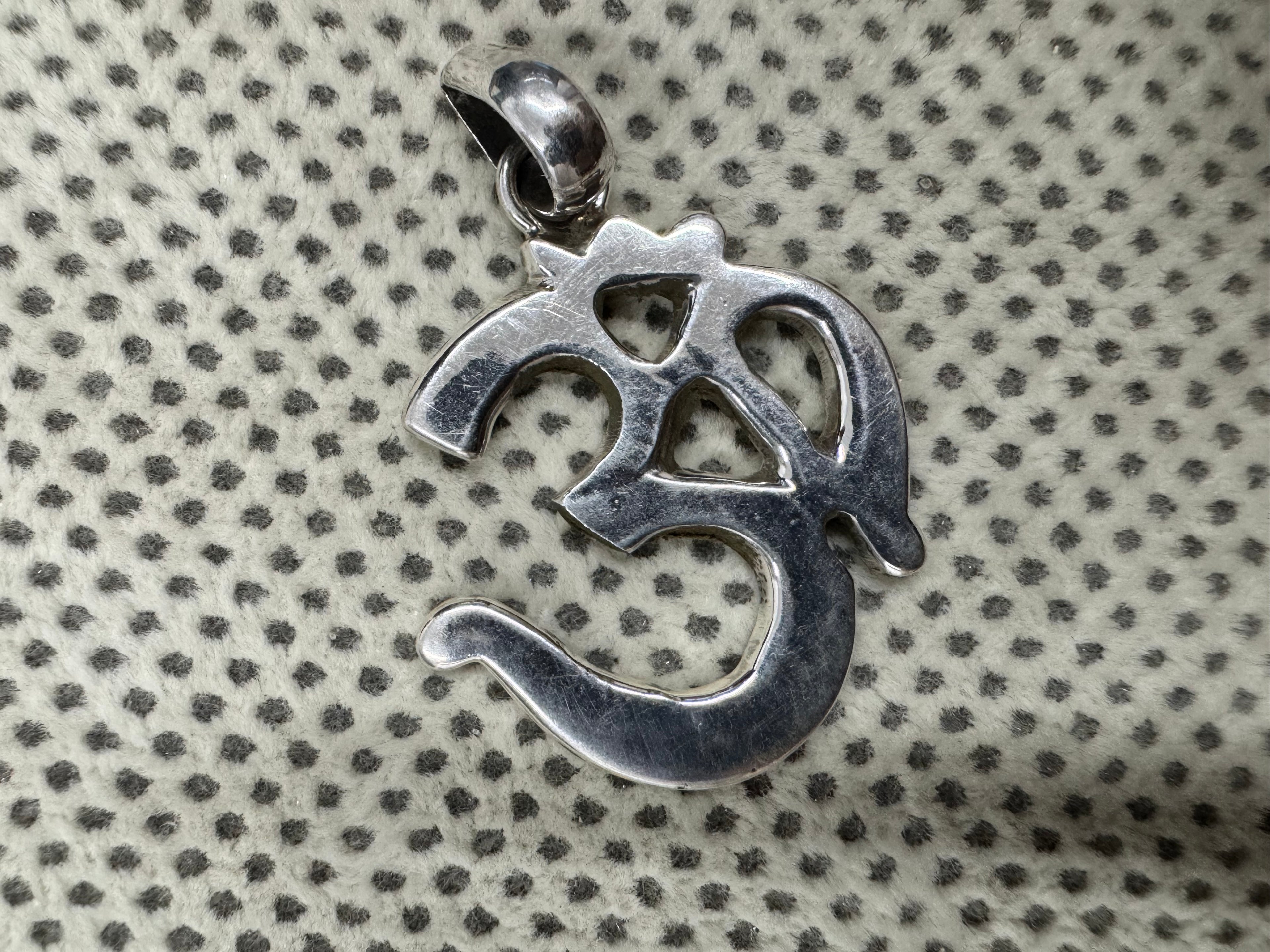 Sterling silver Om pendant photographed face up on dotted cream fabric, showing the polished tapered bail.