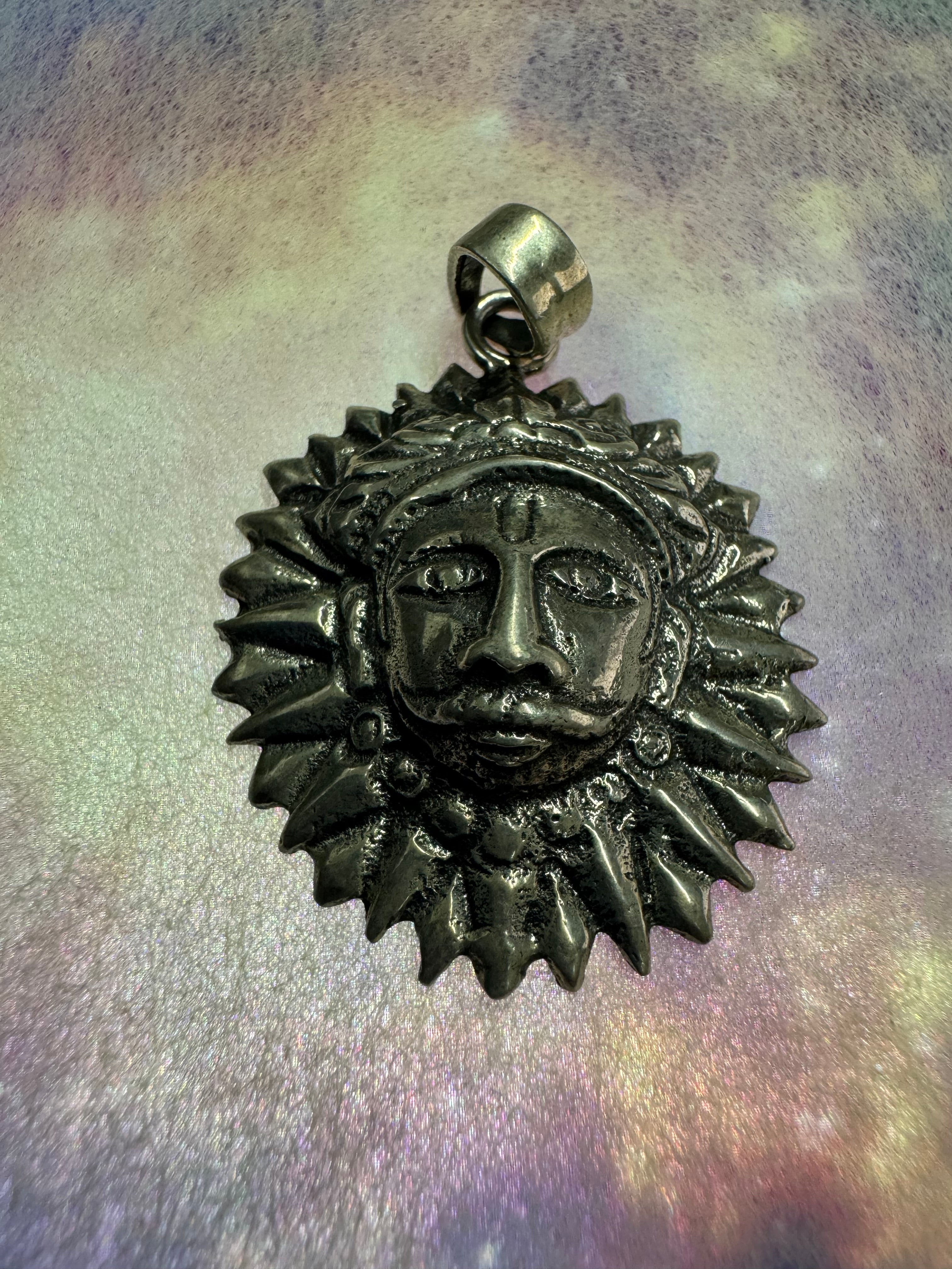 Oxidised sterling silver sun disc pendant with pointed rays around a raised face, on a pale iridescent surface.