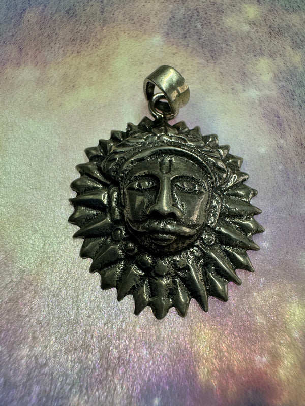 Oxidised sterling silver sun disc pendant with pointed rays around a raised face, on a pale iridescent surface.