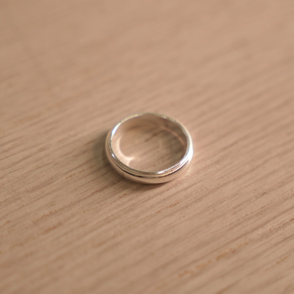 Classic polished sterling silver band ring with milgrain edges, lying on a pale wooden surface.