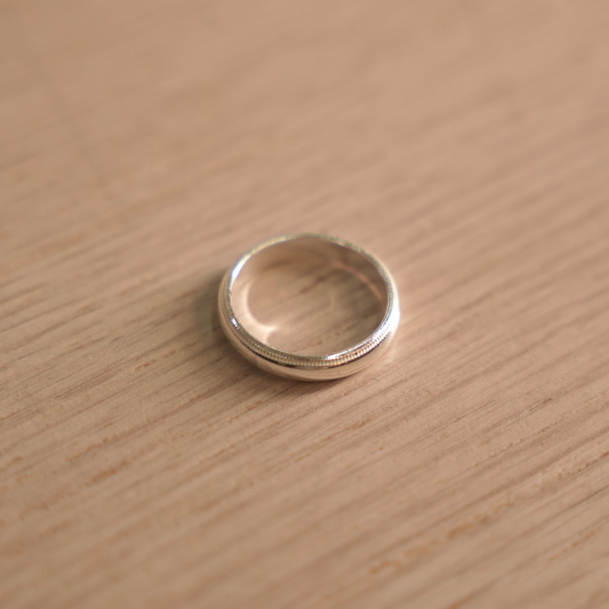 Classic polished sterling silver band ring with milgrain edges, lying on a pale wooden surface.