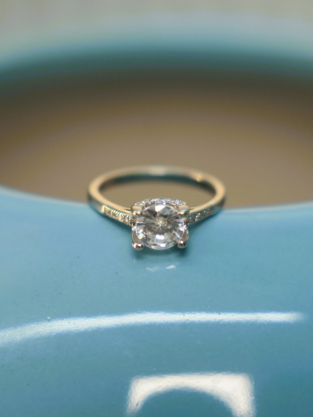 Sterling silver solitaire ring with a round American Diamond in a four prong setting, resting on the rim of a blue vase.
