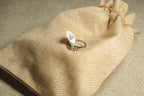 Sterling silver solitaire ring with an oval cubic zirconia, resting on a jute drawstring pouch beside its size tag.