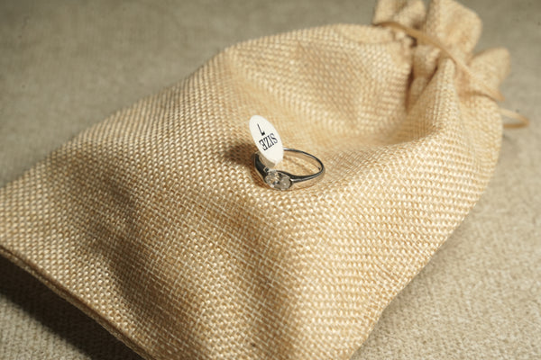 Sterling silver solitaire ring with an oval cubic zirconia, resting on a jute drawstring pouch beside its size tag.