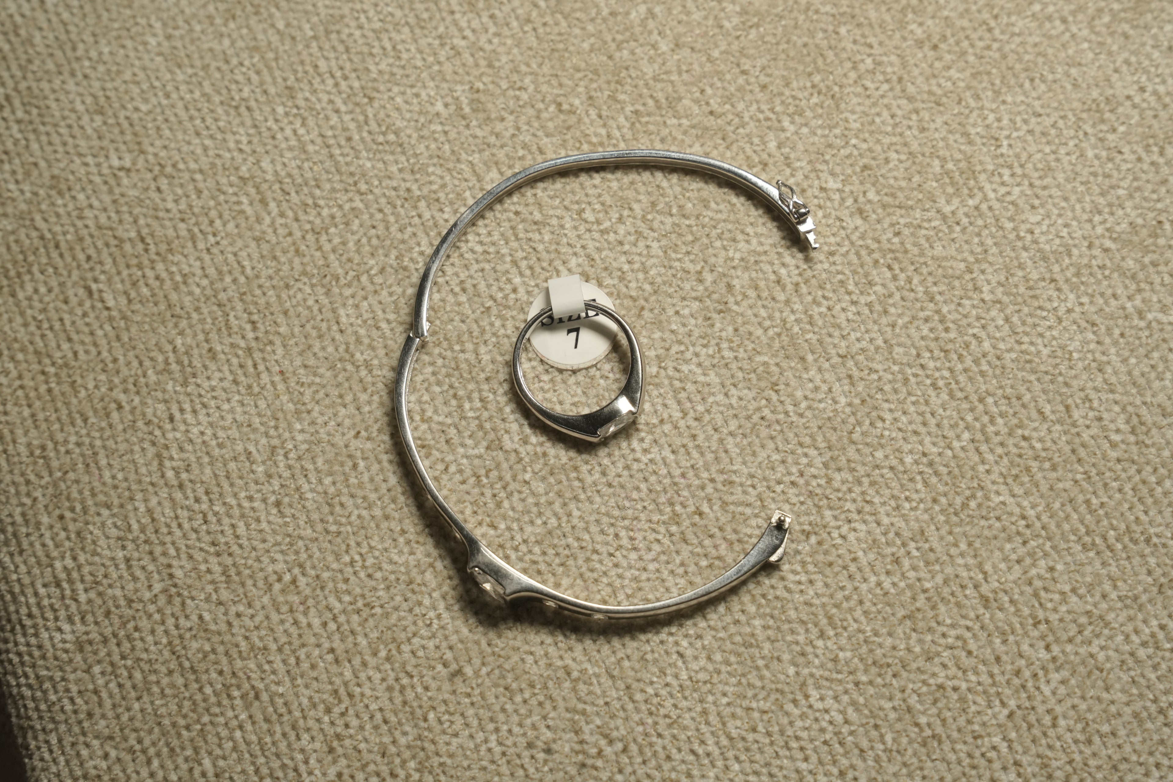 Sterling silver cubic zirconia ring on its size tag, framed by a silver bangle used as a prop on woven fabric.