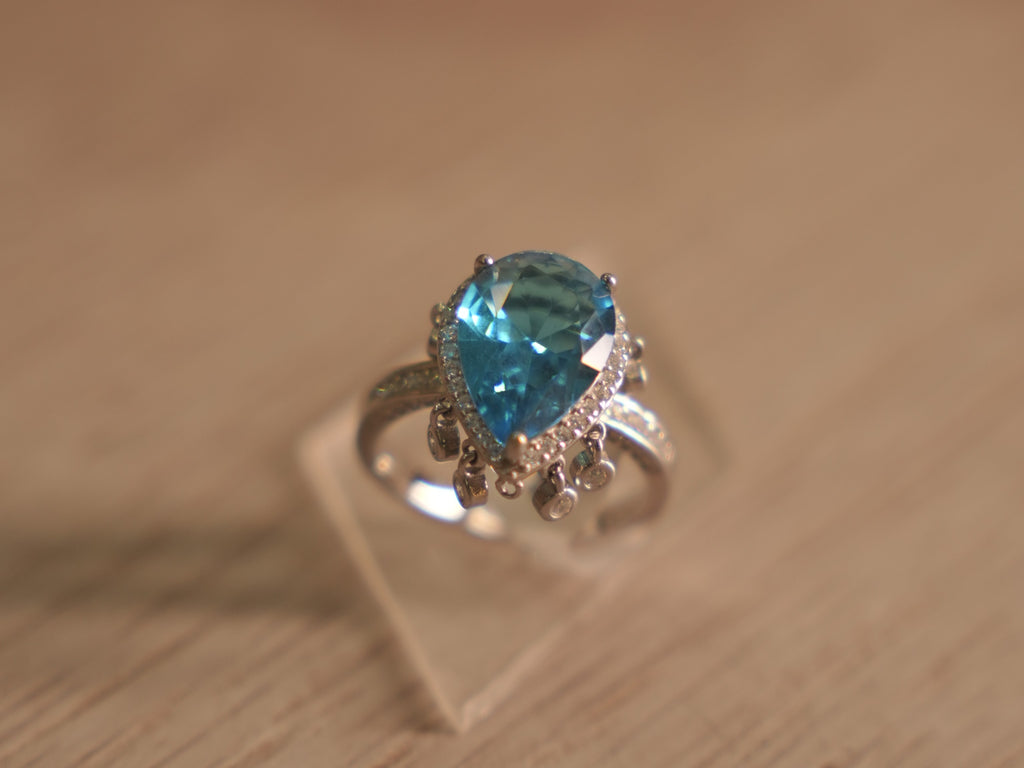 Sterling silver cocktail ring with a pear shaped sky blue glass stone in a halo, with tiny bezel drops below.