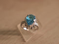 Sterling silver cocktail ring with a pear shaped sky blue glass stone in a halo, with tiny bezel drops below.