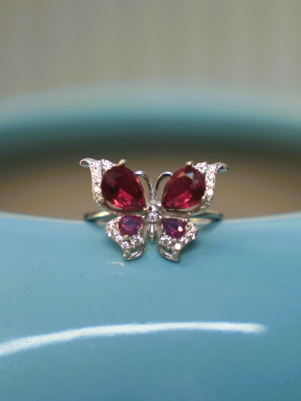 Sterling silver butterfly ring with deep red and pink stone wings, resting on the rim of a blue vase.