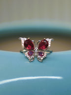 Sterling silver butterfly ring with deep red and pink stone wings, resting on the rim of a blue vase.