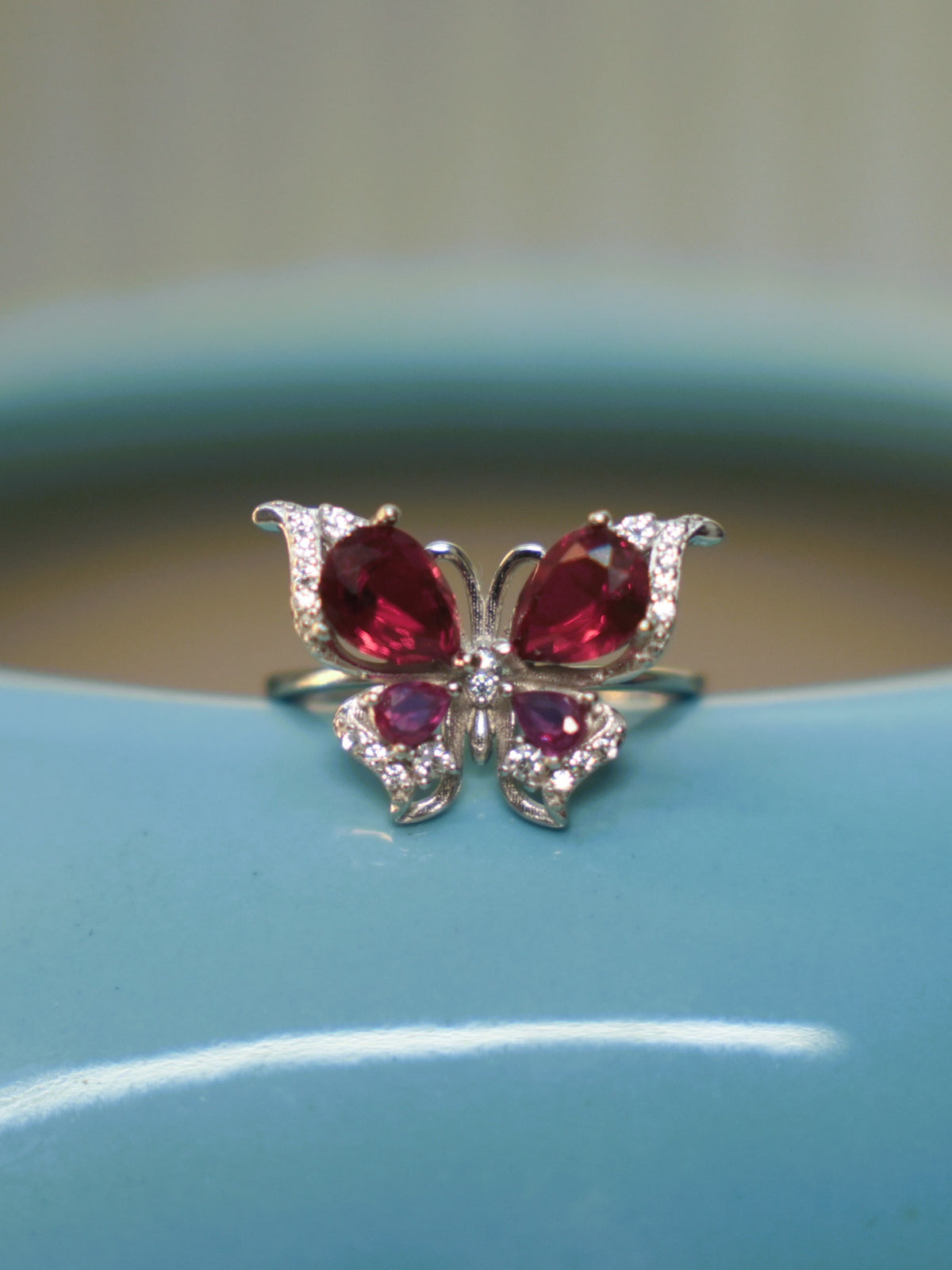 Sterling silver butterfly ring with deep red and pink stone wings, resting on the rim of a blue vase.