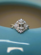 Sterling silver ring with an emerald cut American Diamond in a floral halo of marquise and round stones, on the rim of a blue vase.