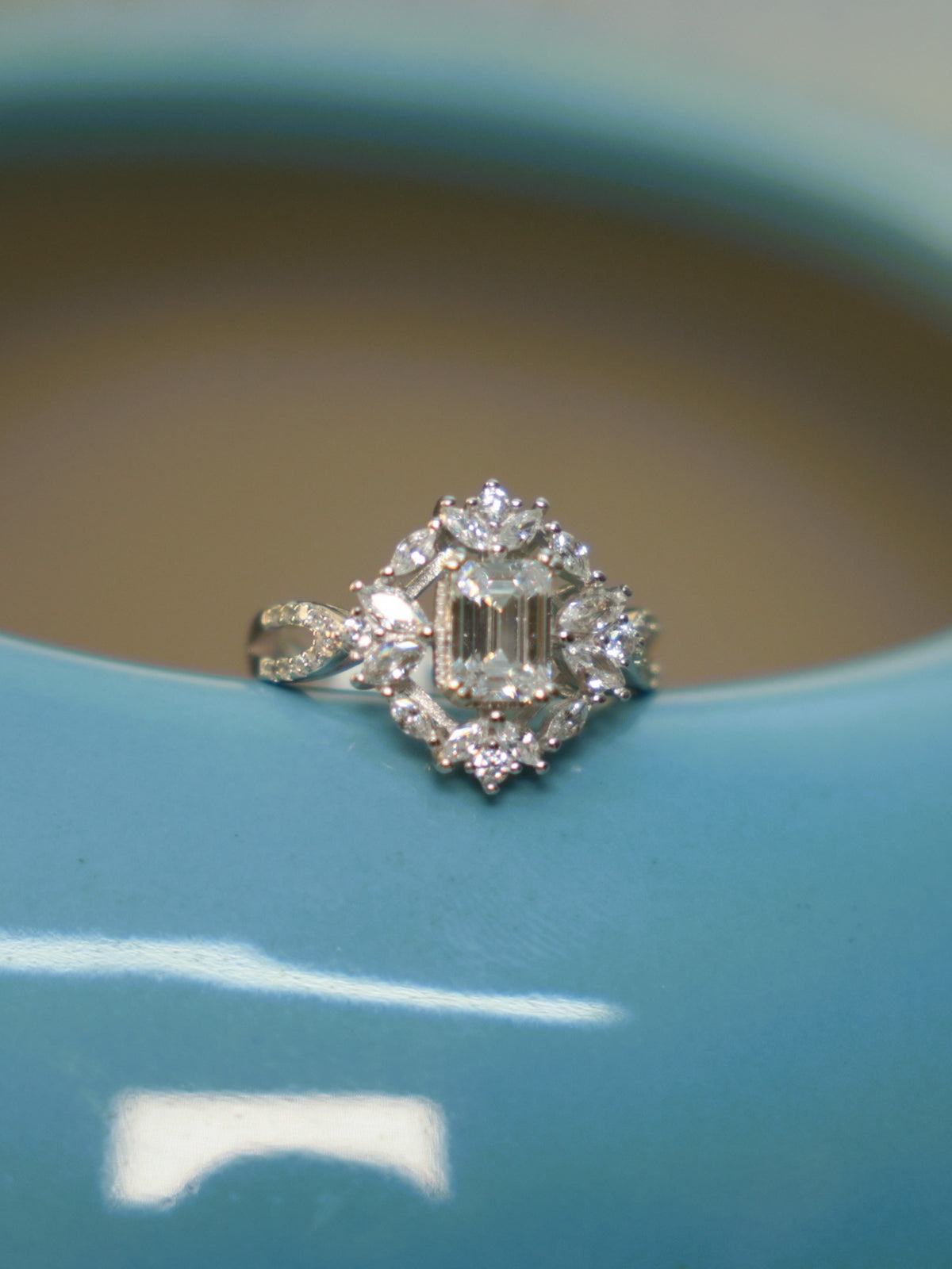 The emerald cut halo ring seen slightly from the side, showing the crossed pave set shoulder.