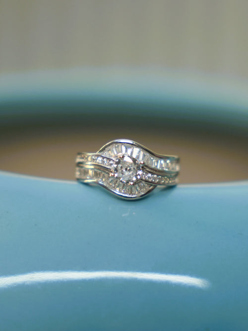 Sterling silver wave ring with a round American Diamond between curved rows of baguette stones, on the rim of a blue vase.